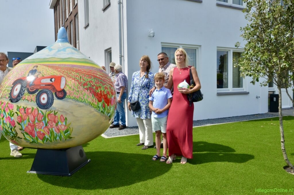 Familie Ruigrok staat buiten bij een groot kunstwerk in de vorm van een bloembol, beschilderd met een bollenveld en tractor. De foto is genomen bij een nieuw gebouw in de Bollenstreek, op kunstgras voor de ingang.