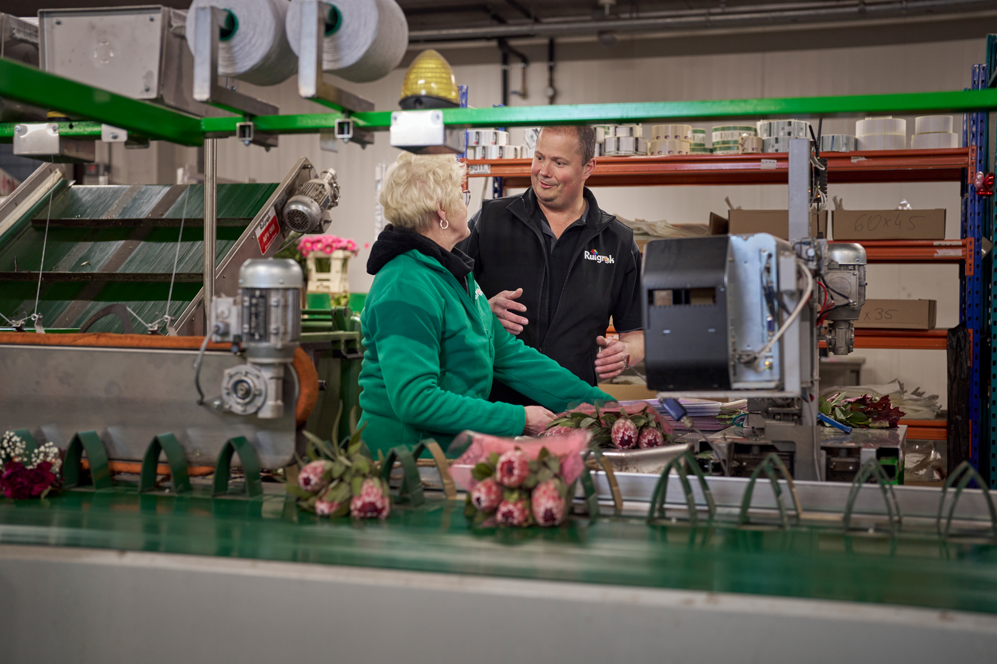 Medewerker en leidinggevende van Ruigrok Groep overleggen bij de lopende band tussen verse bloemen, tijdens het productieproces in de hal