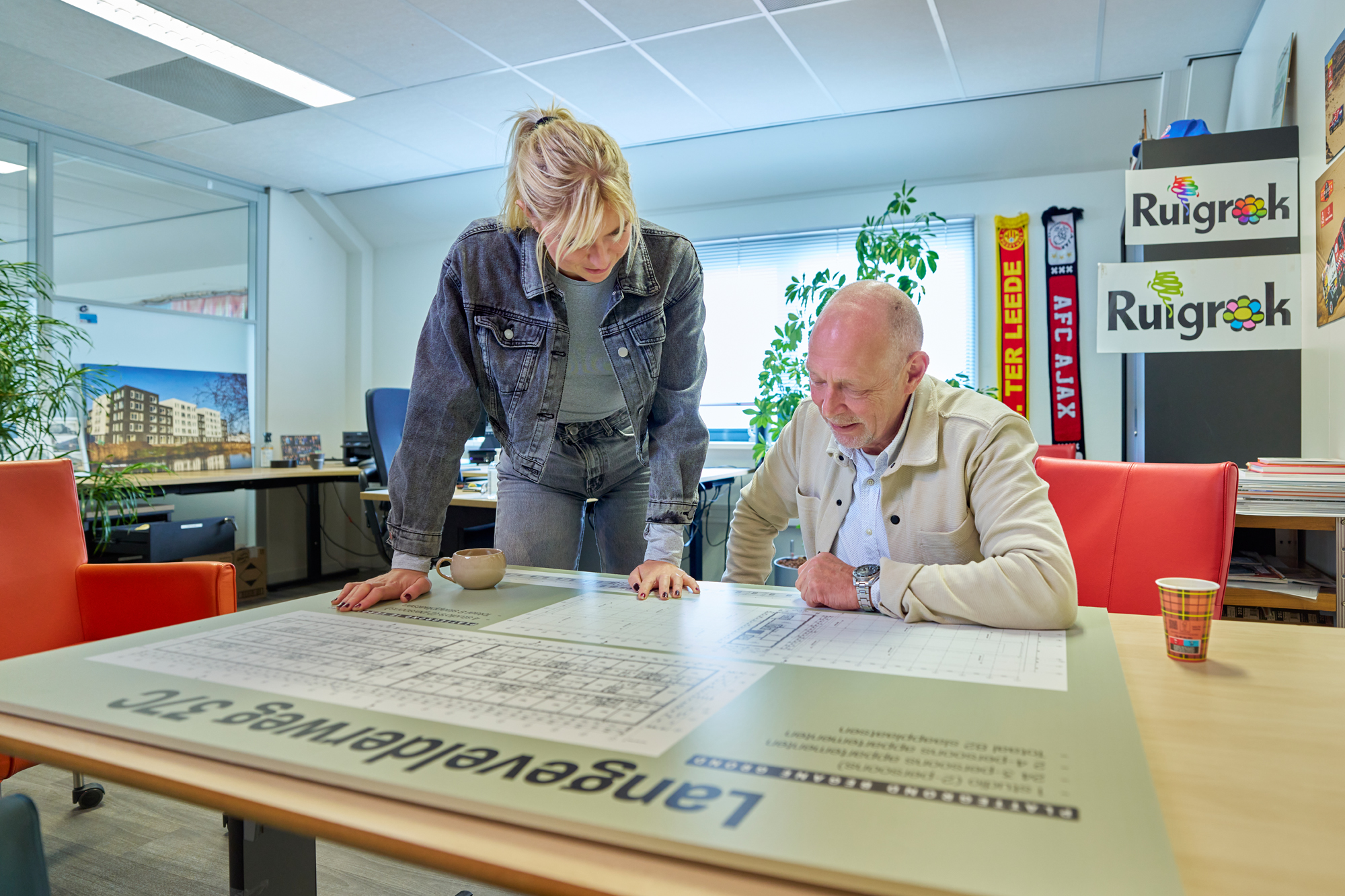 Twee medewerkers van Ruigrok bespreken bouwtekeningen van een huisvestingsproject op kantoor. Op de achtergrond zijn Ruigrok-logo’s en foto’s van woningen te zien