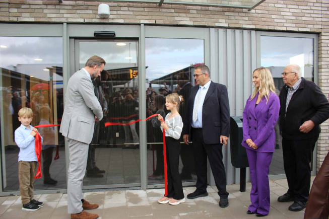 Officiële opening van een nieuw gebouw waarbij een rood lint wordt doorgeknipt door een man in grijs pak en een jong meisje. Naast hen staan meerdere aanwezigen, waaronder vertegenwoordigers van Ruigrok, voor de glazen entree van het pand